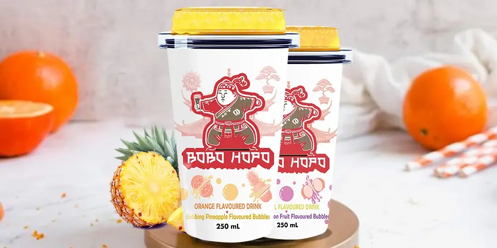 Bobo Hopo flavored bubble drinks in orange and tropical flavours are presented with fresh pineapples and oranges on a textured background.